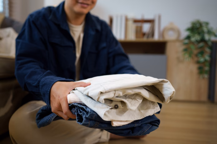 a-smiling-asian-man-holds-a-neat-stack-of-folded-clothing-indoors-ideal-for-concepts-of-decluttering-donation-laundry-and-home-organization-stockpack-istock A smiling Asian man holds a neat stack of folded clothing indoors, ideal for concepts of decluttering, donation, laundry, and home organization.