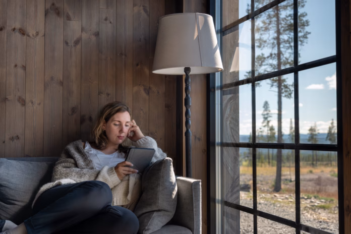 a-woman-relaxes-on-a-sofa-while-reading-on-her-e-reader-in-a-comfortable-wool-vest-enjoying-the-natural-light-streaming-through-the-window-in-a-scenic-cabin-setting-in-a-forest-stockpack-istock A woman relaxes on a sofa while reading on her E-Reader, in a comfortable wool vest enjoying the natural light streaming through the window in a scenic cabin setting in a forest