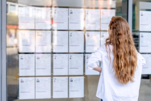 A woman with long hair stands in front of a glass window, carefully reviewing job listings that are displayed on the wall outside a public space.