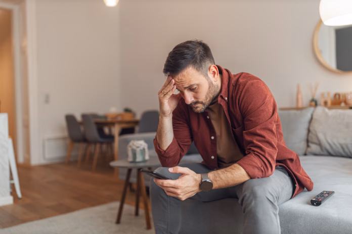 a-worried-adult-man-reading-bad-news-and-text-messages-on-his-phone-while-sitting-on-a-couch-in-his-apartment-stockpack-istock A worried adult man reading bad news and text messages on his phone while sitting on a couch in his apartment.