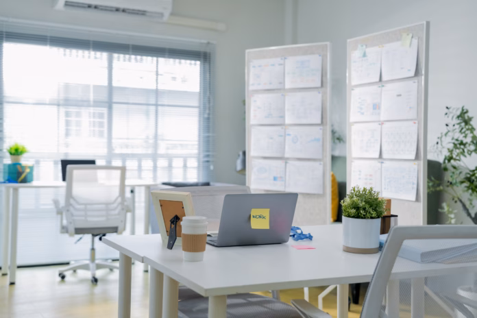 An empty white desk holds a closed laptop with a "work" sticky note, coffee cup, and picture frame, set against a bright office backdrop with wall-mounted planning charts — a clean, minimalist workspace scene.