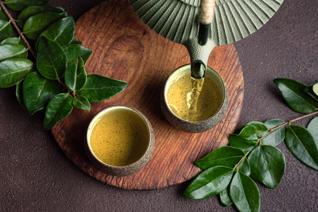 Asian iron teapot pouring green tea into cups close up. Traditional japanese chinese tea set on dark brown background with green leaves, top view. Aesthetic asian tea ceremony.