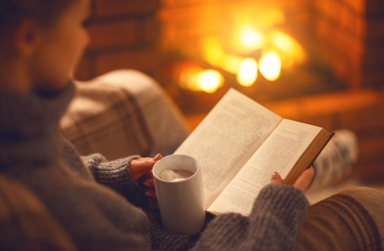 book and cup of coffee in hands of girl on winter autumn evening near fireplace