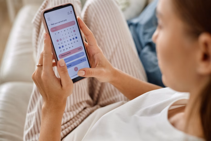 caucasian-young-adult-woman-holding-smartphone-while-checking-menstrual-cycle-calendar-app-sitting-on-sofa-with-striped-pants-visible-focusing-on-period-tracking-application-interface-stockpack-istock Caucasian young adult woman holding smartphone while checking menstrual cycle calendar app, sitting on sofa with striped pants visible, focusing on period tracking application interface
