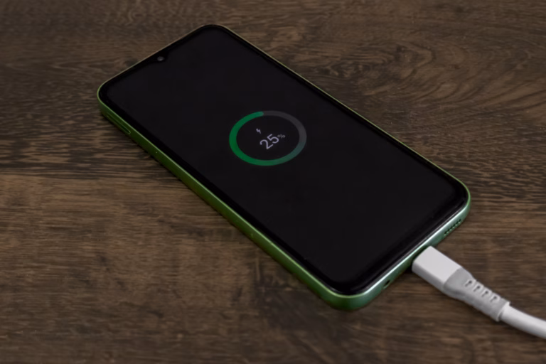 Green smartphone charging via USB cable on a wooden table, showing 25% battery. Concept of mobile technology, low power, energy use, connectivity and digital routine.