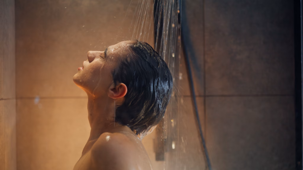 Portrait sexy woman taking shower alone. Closeup nude young model washing hair standing in soft light bathroom. Closed eyes pretty brunette enjoying hot water drops in bath. Self love moment concept