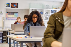 Serious multiethnic girl student wearing eyeglasses while working on laptop in a classroom with copy space. Focused african american girl typing on laptop with study material on desk. Guys and girls using laptop during computer lesson in secondary classroom.