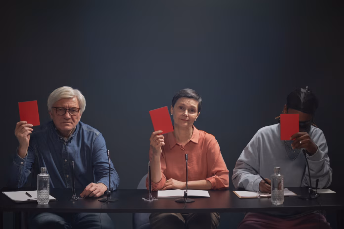 three-diverse-judges-sitting-at-table-holding-up-red-cards-during-talent-competition-including-caucasian-man-caucasian-woman-and-black-man-microphones-and-notepads-visible-stockpack-istock Three diverse judges sitting at table holding up red cards during talent competition, including Caucasian man, Caucasian woman and Black man, microphones and notepads visible