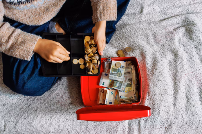 top-view-of-a-young-woman-sitting-on-a-bed-while-counting-euro-coins-in-an-open-red-cash-box-filled-with-euro-banknotes-stockpack-istock Top view of a young woman sitting on a bed while counting euro coins in an open red cash box filled with euro banknotes