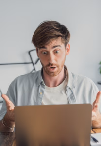 Young man sitting at his desk reacting with a shocked and bewildered expression as he looks at unexpected bad news or a surprising message on his laptop while working from home