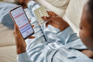 Black woman holding smartphone displaying menstrual cycle tracking app while examining blister pack of birth control pills, sitting on couch in relaxed position, focusing on reproductive health