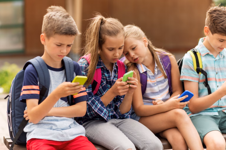 primary education, friendship, childhood, technology and people concept - group of happy elementary school students with smartphones and backpacks sitting on bench outdoors