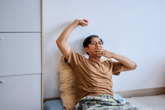 sleepy-asian-man-stretching-his-arm-and-yawning-while-sitting-up-in-bed-at-home-stockpack-istock Sleepy Asian Man Stretching His Arm and Yawning While Sitting Up in Bed at Home