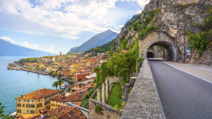 View of the road hugging the rocky cliff face above Lake Garda, featuring a stone tunnel entrance. The picturesque village of Limone sul Garda is visible below, Lombardy, Italy