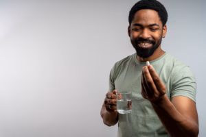 Young black man taking pill with glass of water, isolated on clean background, health and wellness concept